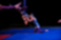Two students in galaxy print unitard laugh together while performing. One hangs upside down on an aerial sling, locked in place by her knee across the sling's fabric, while the other kneels on the gound and giggles.