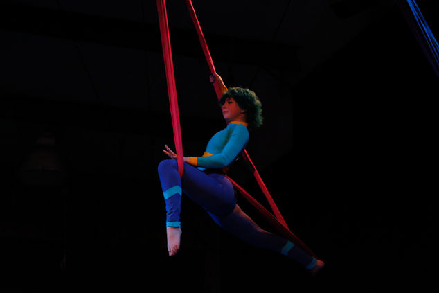 A blue haired female bodied teenager performs on a red aerial sling. 