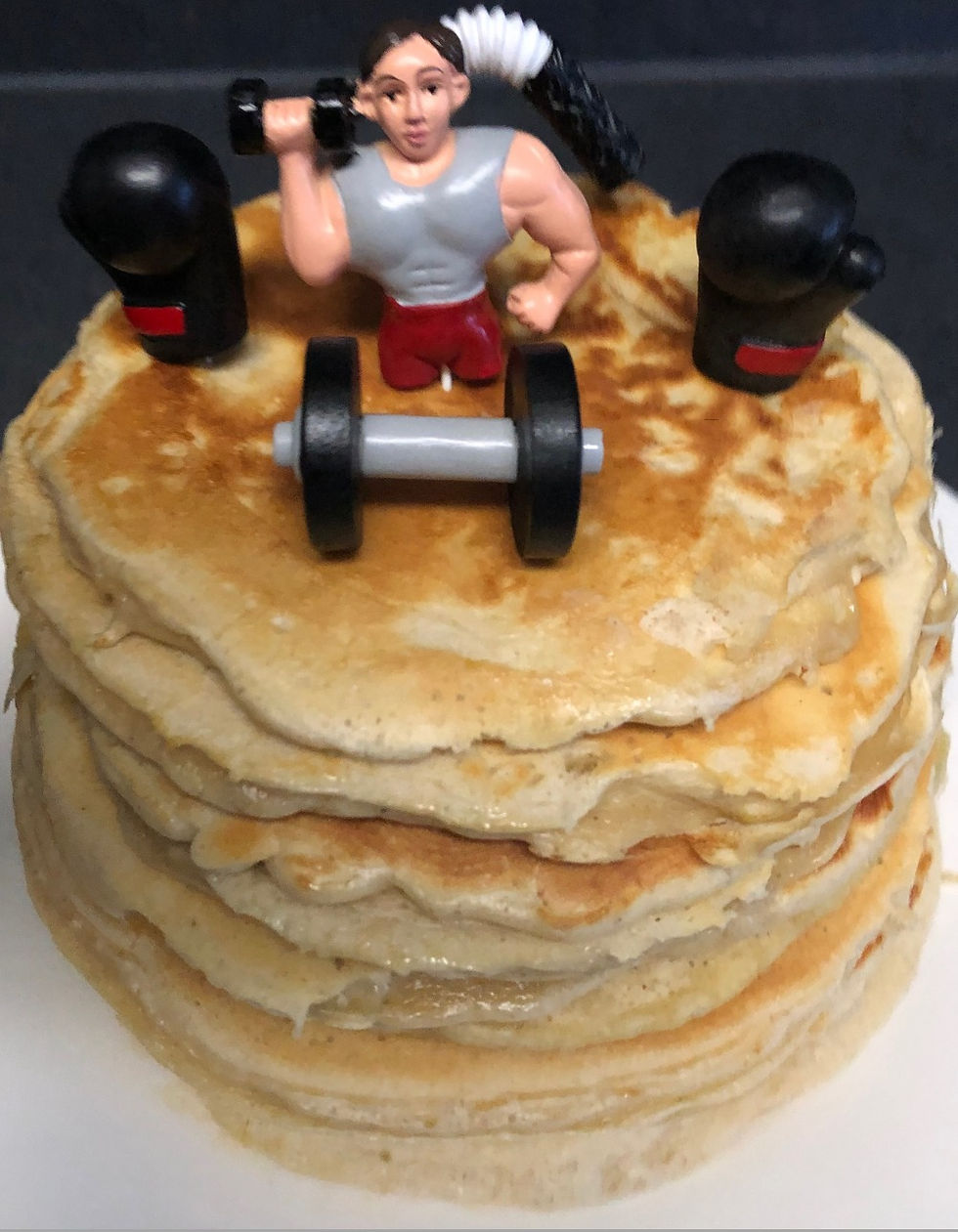 Fitnessturm bestehend aus übereinander gestapelten Proteinpacakes. Oben auf sitzt der Tortenaufsatz, bestehend aus zwei schwarzen Boxhandschuhen, Gewichtheber, der eine Hantel hochhebt mit dem rechten Arm, eine Kurzhantel und eine Biegehantel