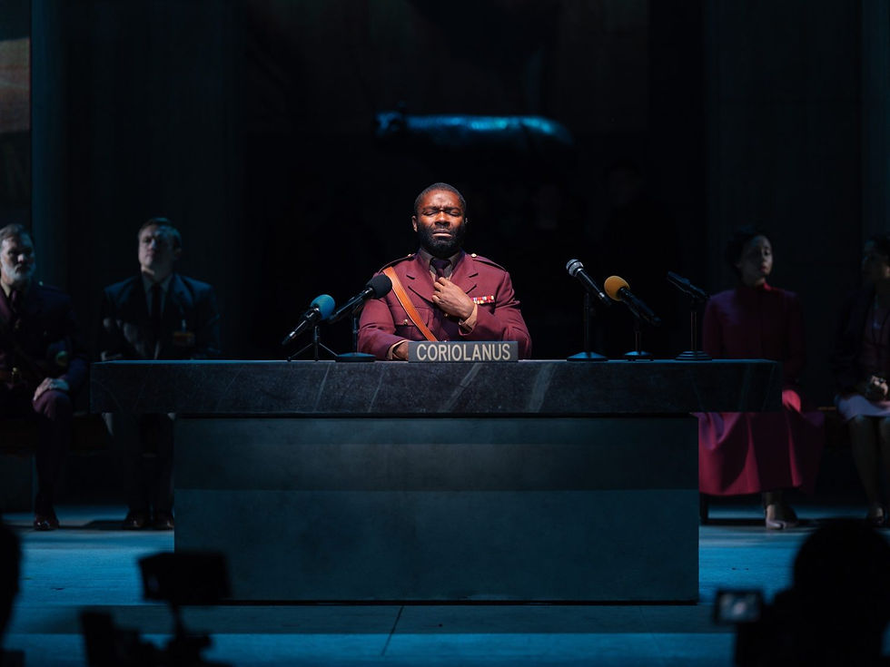 David Oyelowo as Coriolanus at the National Theatre. A man on stage speaking to the press, with a nametag on a desk that reads 'Coriolanus' and multiple microphones pointed at the man.