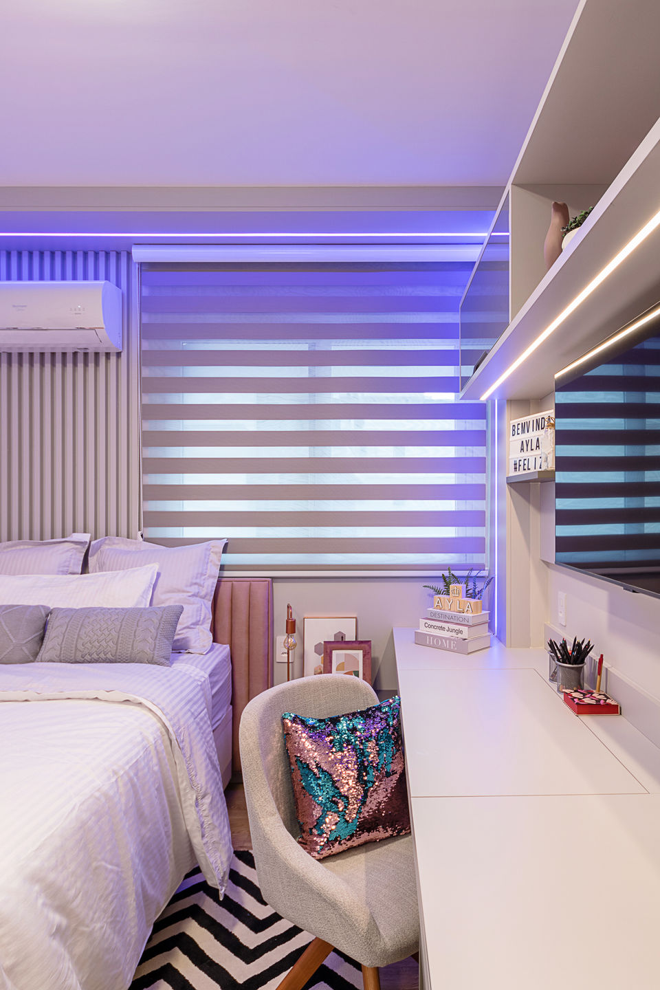 Quarto feminino com cama de casal, iluminação LED RGB embutida, escrivaninha branca com prateleiras, cadeira estofada com almofada metalizada e persiana horizontal dupla. Decoração jovem e contemporânea com tapete geométrico preto e branco e painel ripado atrás da cama.