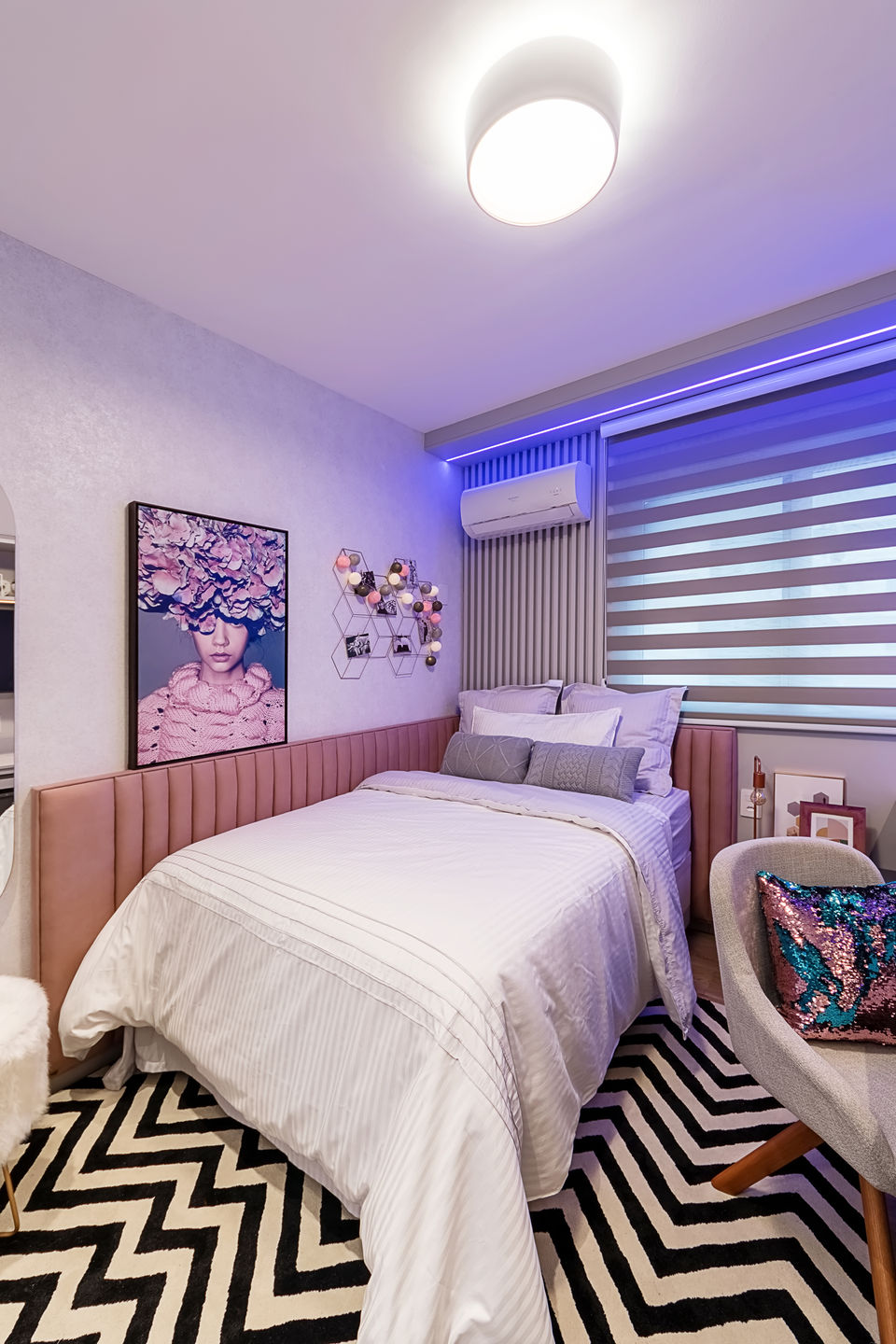 Quarto feminino com cama de casal, iluminação LED RGB embutida, escrivaninha branca com prateleiras, cadeira estofada com almofada metalizada e persiana horizontal dupla. Decoração jovem e contemporânea com tapete geométrico preto e branco e painel ripado atrás da cama.