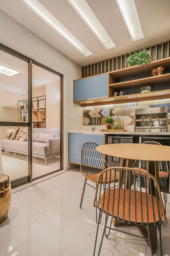 Varanda gourmet com marcenaria azul e madeira clara, revestimento geométrico, mesa redonda com cadeiras em metal e acesso integrado à sala de estar. Projeto contemporâneo e funcional em apartamento de alto padrão.