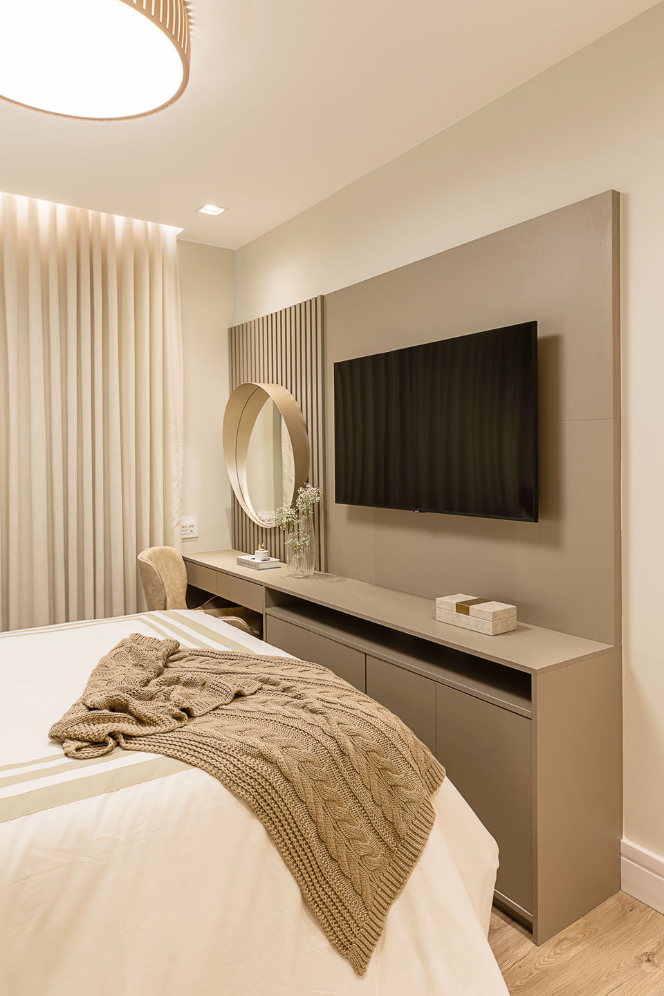 Quarto de casal com paleta neutra, cama com enxoval bege e cabeceira estofada, criados-mudos com luminárias, cortinas de linho até o piso, TV fixada na parede e penteadeira com espelho redondo iluminado. Iluminação central com plafon de design contemporâneo e atmosfera acolhedora.