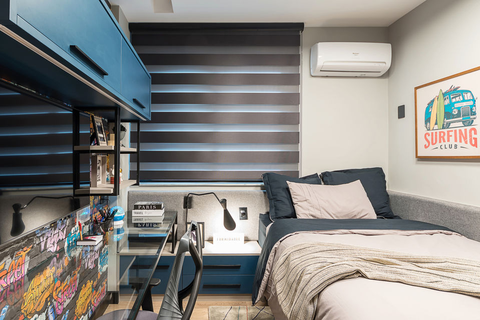 Quarto juvenil com cama de solteiro, marcenaria azul e cinza, escrivaninha com cadeira acrílica e iluminação de tarefa. Persiana horizontal dupla, ar-condicionado e decoração com pôsteres e objetos personalizados. Projeto funcional com layout compacto e estilo contemporâneo.