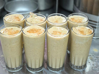 Eight glasses filled with creamy, frothy lassi topped with crushed ice on a steel counter, creating a refreshing, inviting look. Madurai's famous jigarthanda.