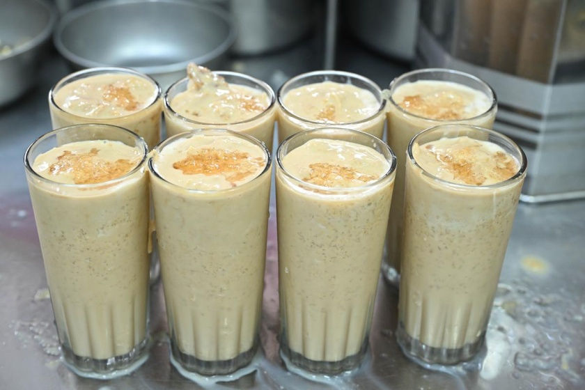 Eight glasses filled with creamy, frothy lassi topped with crushed ice on a steel counter, creating a refreshing, inviting look.  Madurai's famous jigarthanda.