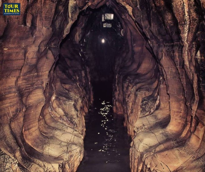 A narrow, rocky cave with textured walls and a river running through it. Dim lighting creates a mysterious atmosphere. "Tour Times" text is visible.Ramayana Yatra Tour