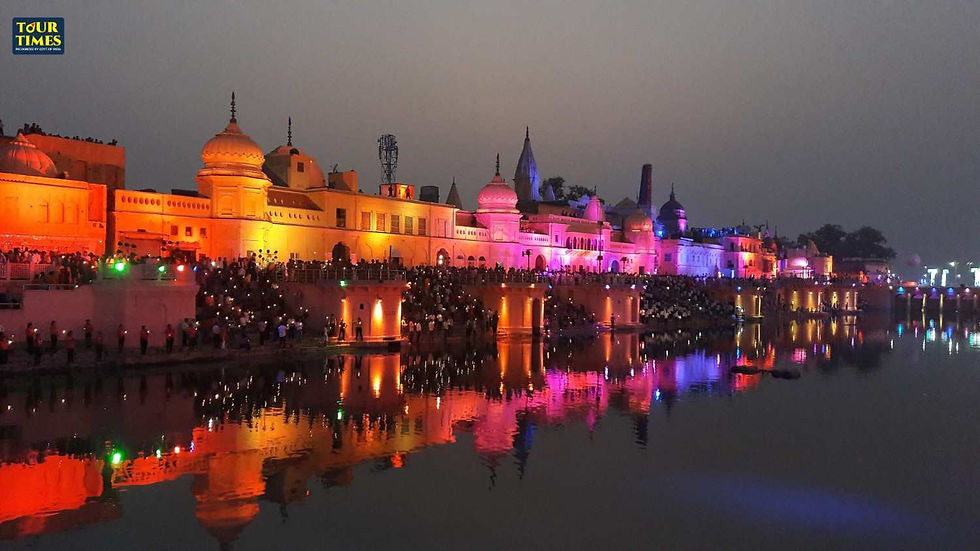 Crowds gather near a brightly lit temple at dusk with colorful lights reflecting on the water. A "Tour Times" sign is visible above.Bharat Darshan Yatra