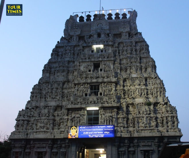 Intricately carved temple facade at dusk, illuminated against a blue sky. A blue sign with Tamil text is visible at the entrance. Mood: serene.Thiru Kalvanoor Perumal Temple