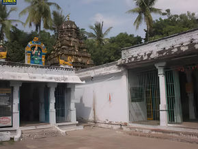 Temple entrance with ornate figures and tropical trees in the background. Weathered white walls, barred gates, and "TOUR TIMES" text.Pachai Vanna Perumal Temple a