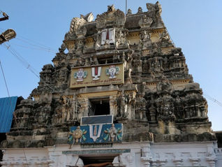 Ancient temple facade with intricate stone carvings, banners with symbols and text. Sunny day, blue sky. "Tour Times" logo in corner.Ashtabuja Perumal Temple