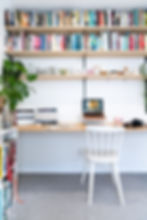 A home office with a wooden desk, laptop, camera, and cookbooks. White chair with book shelves above