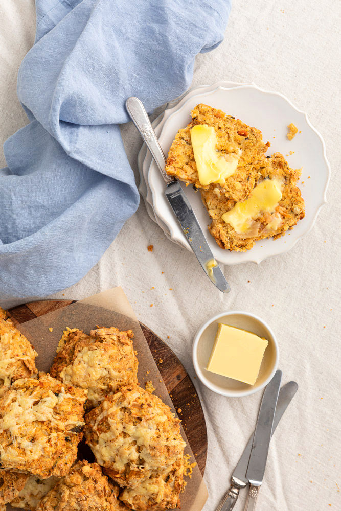 Cheesy scones on a wooden board with a pat of butter and a knife. A scone is on a plate, sliced open with melted butter.