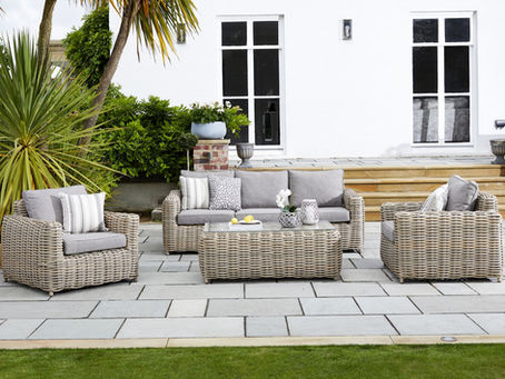 Wicker patio furniture with gray cushions on a stone patio, surrounded by lush greenery. A white house with large windows is in the background.
