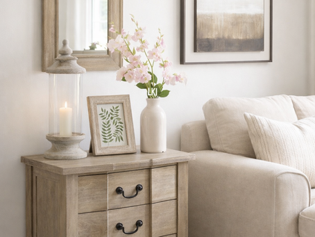 Beige living room with a wooden table, pink flowers, a candle, and framed art. Cozy vibe with a mirror and abstract painting on the wall.