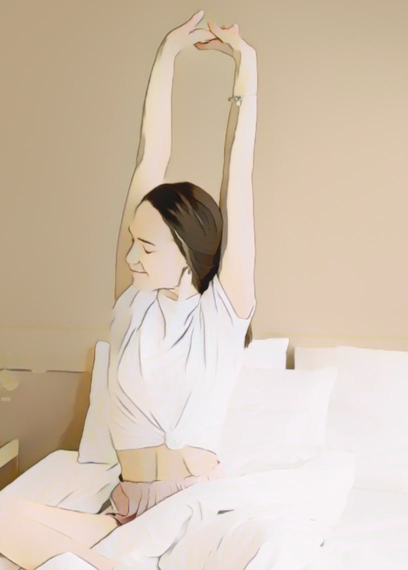 Woman sitting on a bed, stretching with arms up, eyes closed, smiling. Wearing a white top and pink shorts. Soft, neutral-toned room.