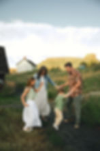 Family of four holding hands and dancing on a grassy path with house and butte in background. Casual summer attire, joyful mood.