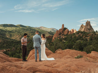 Bride and groom exchange vows on red rocks as officiant reads, with green mountains and dramatic sandstone spires under blue sky.