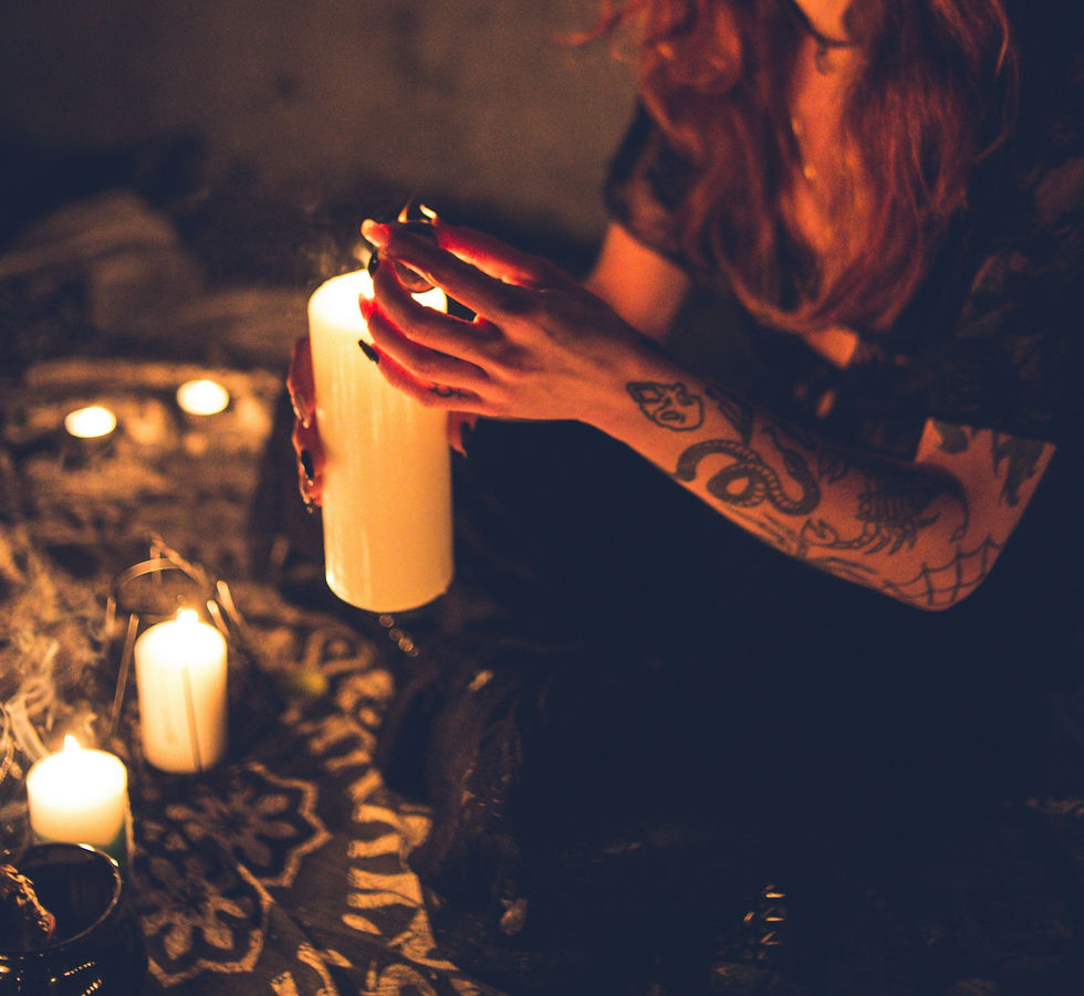 A woman with arm tattoos holding a white candle and sitting cross-legged on the floor in candlelight.