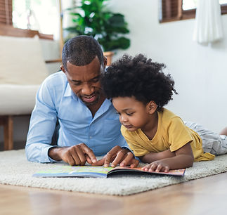 A father and child are lying on a carpeted floor, reading a book together