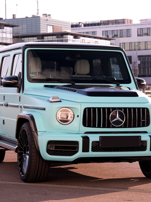 Mercedes-Benz G-Klasse AMG in Pastell-Türkis auf einem städtischen Parkplatz, im Hintergrund moderne Bürogebäude – markanter Luxus-SUV mit sportlicher Präsenz und urbanem Flair.
