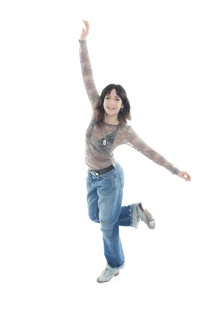 Photograph of a girl wearing jeans, a gray lace blouse, and tap shoes, against a white background, with one leg raised behind and arms lifted, smiling.