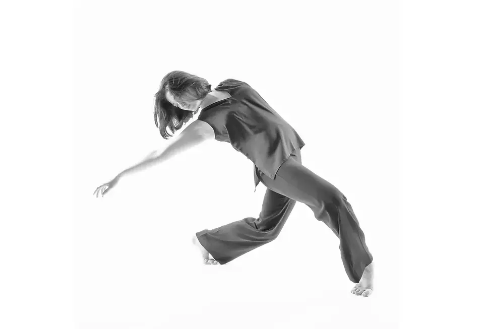 Black and white photograph of a dancer in flowing attire, in motion, with bent legs and arm to the side.