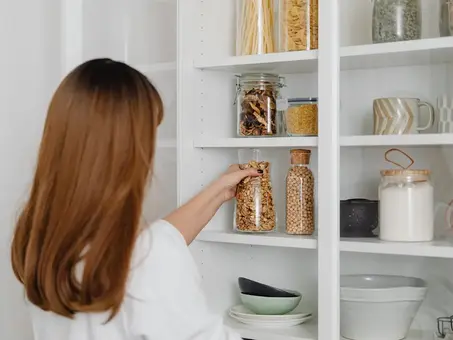 A professional organizer in a white shirt organizing a jar of cereal in a white kitchen cabinet filled with jars of pasta, grains, and mugs.