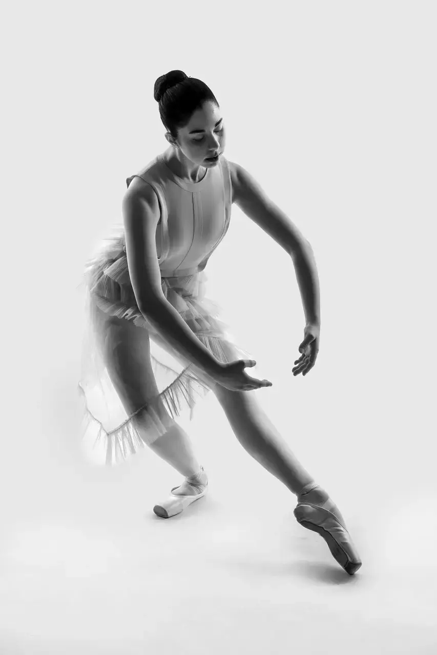 Black and white photo of a classical ballerina wearing a tulle dress and pointe shoes, stretching one leg forward while the other is in plié, and arms extended in front, in first position.