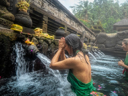 Women in green sarongs perform a ritual in a stone pool with water spouts. Tropical setting with offerings placed on the spouts.