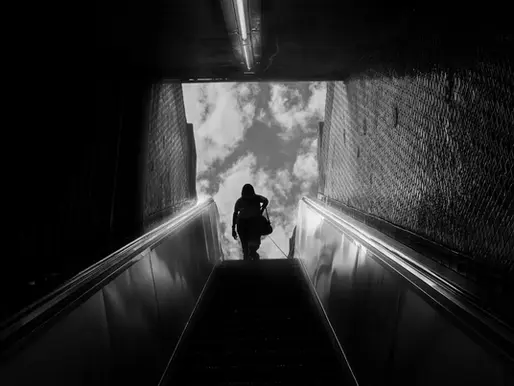 Silhouette of a person ascending an escalator towards a cloudy sky, framed by dark walls. Dramatic contrast in black and white.