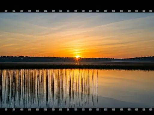 Sunset over a tranquil lake with tall grass reflections. Orange and blue sky creates a peaceful mood. Filmstrip border surrounds the view.
