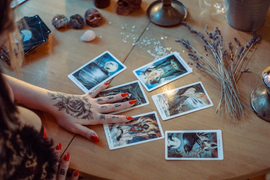 A tattooed hand with red nails spreads tarot cards on a wooden table. Nearby are crystals, skulls, and lavender, creating a mystical vibe.