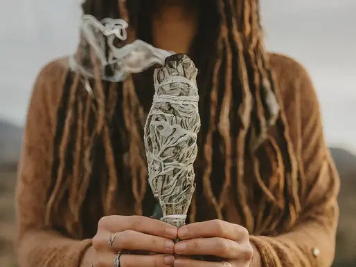 Person with long hair holds a smoking sage bundle outdoors, wearing rings and a brown garment. Calm setting with a blurred background.