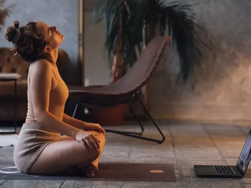 Woman in a beige outfit doing yoga on a mat in a cozy room with a laptop in front of her, looking calm and focused.