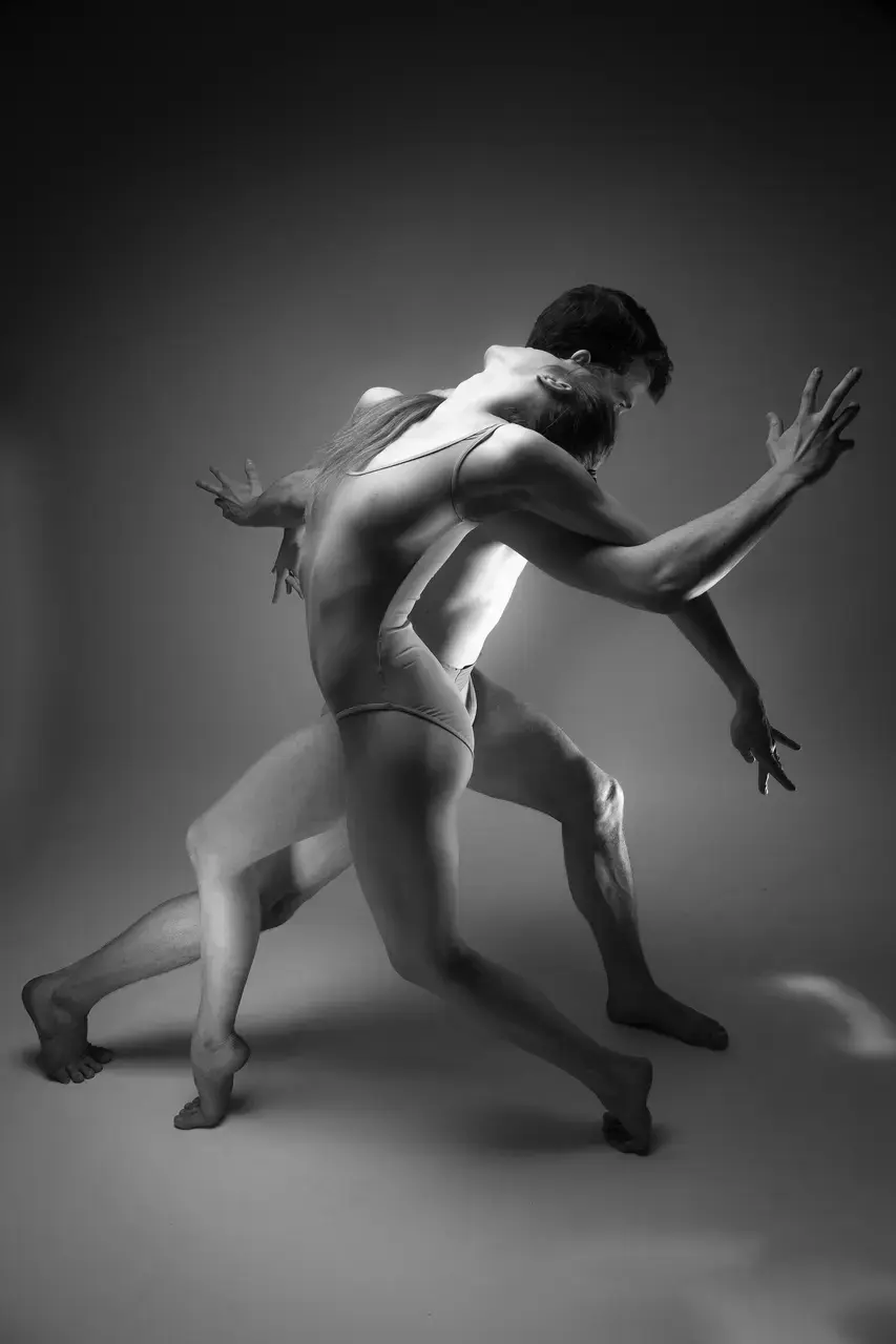 Black and white photograph of a couple of dancers wearing leotards, standing with bent arms and legs, embracing each other from behind.