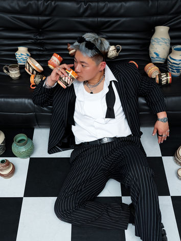 An image of a person sitting on a checkered floor posing in a business professional outfit with their ceramics products