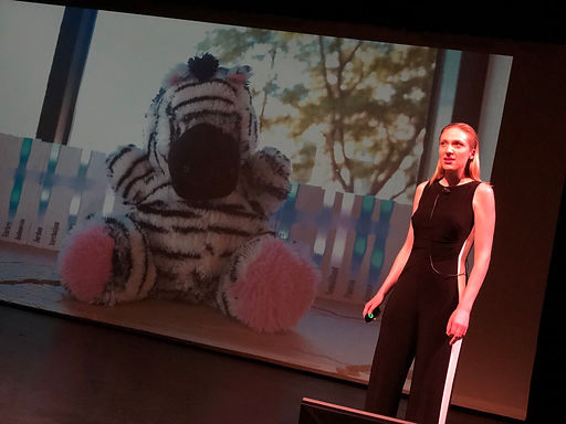 A photo of Layne Jackson Hubbard pitching MindScribe.org at Catalyze CU's Demo Day. She is standing on a stage with a large projector screen behind. The screen displays a stuffed animal zebra robot.