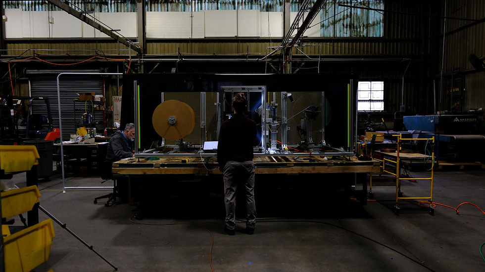 Two people inspect a large industrial machine in a dim workshop, with cables, metal frames, and bright circular disks.