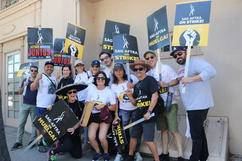 A group shot of Latino actors wearing protest t-shirts and carrying signs. Behind them is a concrete building.
