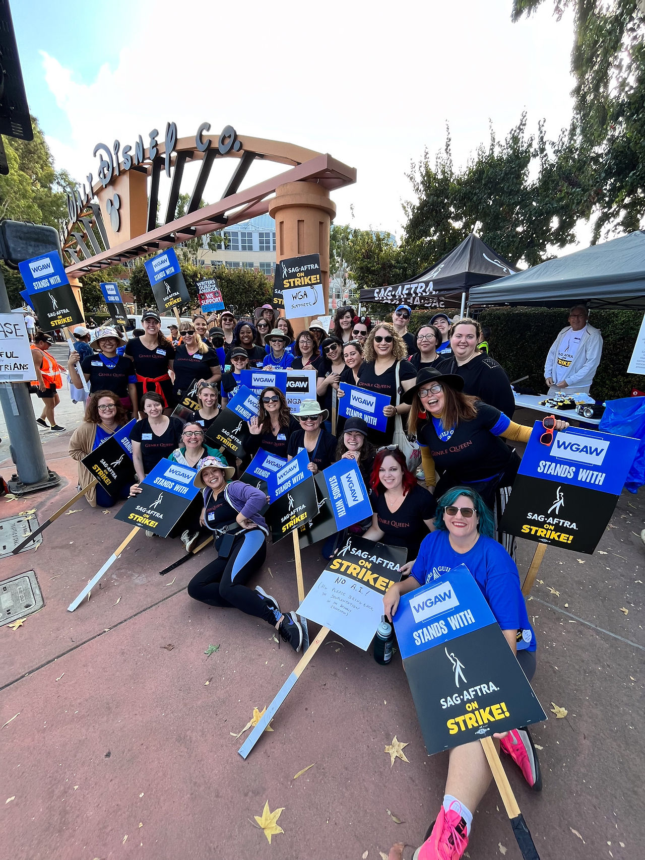A group photo of picketers with WGAW/SAG-AFTRA picket signs take a photo in front of the Walt Disney Studio front gate. A few dozen individuals within the diverse crowd stand in a few lines, with those in the front seated on the ground. Many hold WGAW/SAG-AFTRA picket signs. Seen in the background is the SAG-AFTRA tent with a black tarp bearing the union symbol and copy and a studio gate and sign with "Walt Disney Co." in black Disney-styled copy and the Mickey Mouse silhouette.