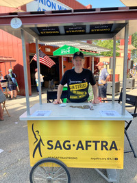 A man wearing a SAG-AFTRA On Strike t-shirt stands behind a booth and looks at the camera. On the front of the booth is a yellow SAG-AFTRA banner.