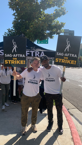 Shea Whigham, left, and an African American man gives each other a side hug as they hold picket signs in their free hand. They both wear white "SAG-AFTRA Strong" t-shirts. Behind them is a SAG-AFTRA tent.  