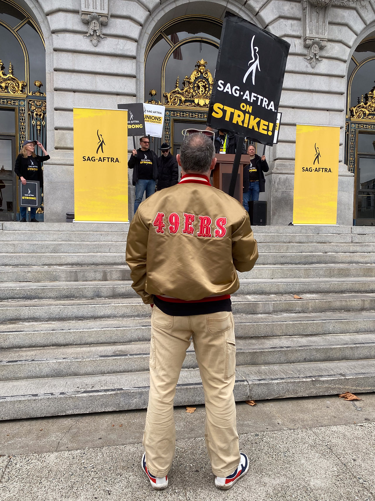 A San Francisco-Northern California Local member listening on to a rally speaker in front of the San Francisco City Hall steps. The member, a man, stands in the center foreground of the photo with his back turned towards the photographer. He wears a gold-colored letterman jacket with "49ers" across the back. The letters have been sewn on; they are written in all-caps and are red in color. Underneath his jacket is a black t-shirt, and he wears khaki pants. In his hand is a "SAG-AFTRA On Strike!" picket sign. In the background are the front steps of SF City Hall. On the landing, right before the front doors, is a podium. On either side are two free-standing SAG-AFTRA banners in yellow bearing the SAG-AFTRA logo and symbol. Some individuals in dark clothing are visible; each holds a SAG-AFTRA Strike picket sign.