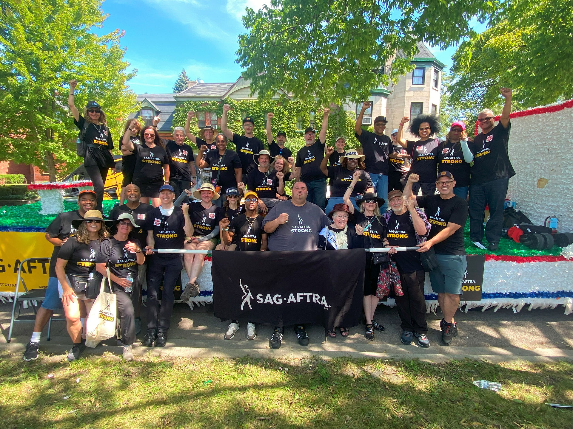 A group shot of Chicago Local members atop their Labor Day Parade float. All wear "SAG-AFTRA Strong" t-shirts and raise their fists. Two individuals in the front row carry a black SAG-AFTRA banner. 