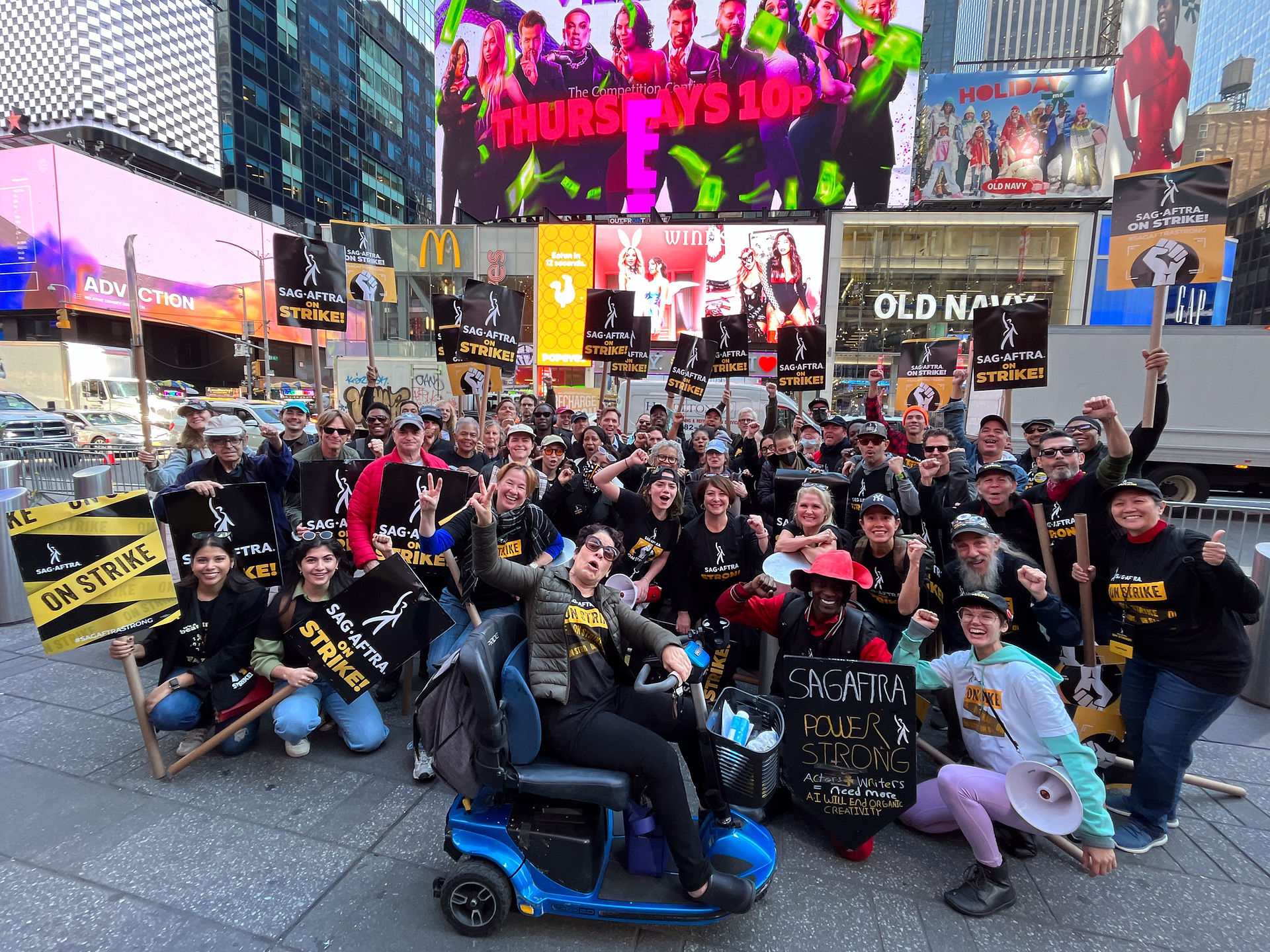 A group shot of New York Local members on the sidewalk in Times Square. Members of the diverse crowd stand in a few row, with those in the front kneeling on the sidewalk. Featured prominently, center front, is member Christine Bruno, who is seated in her electric wheelchair. Many members wear SAG-AFTRA Strike t-shirts; signs seen throughout. Behind them, on the other side of the street, are store fronts and billboard signs. Traffic is also visible.
