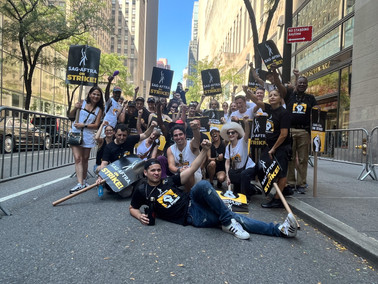 A group photo of protesters on a street. Many wear SAG-AFTRA Strike t-shirts. Signs seen throughout.