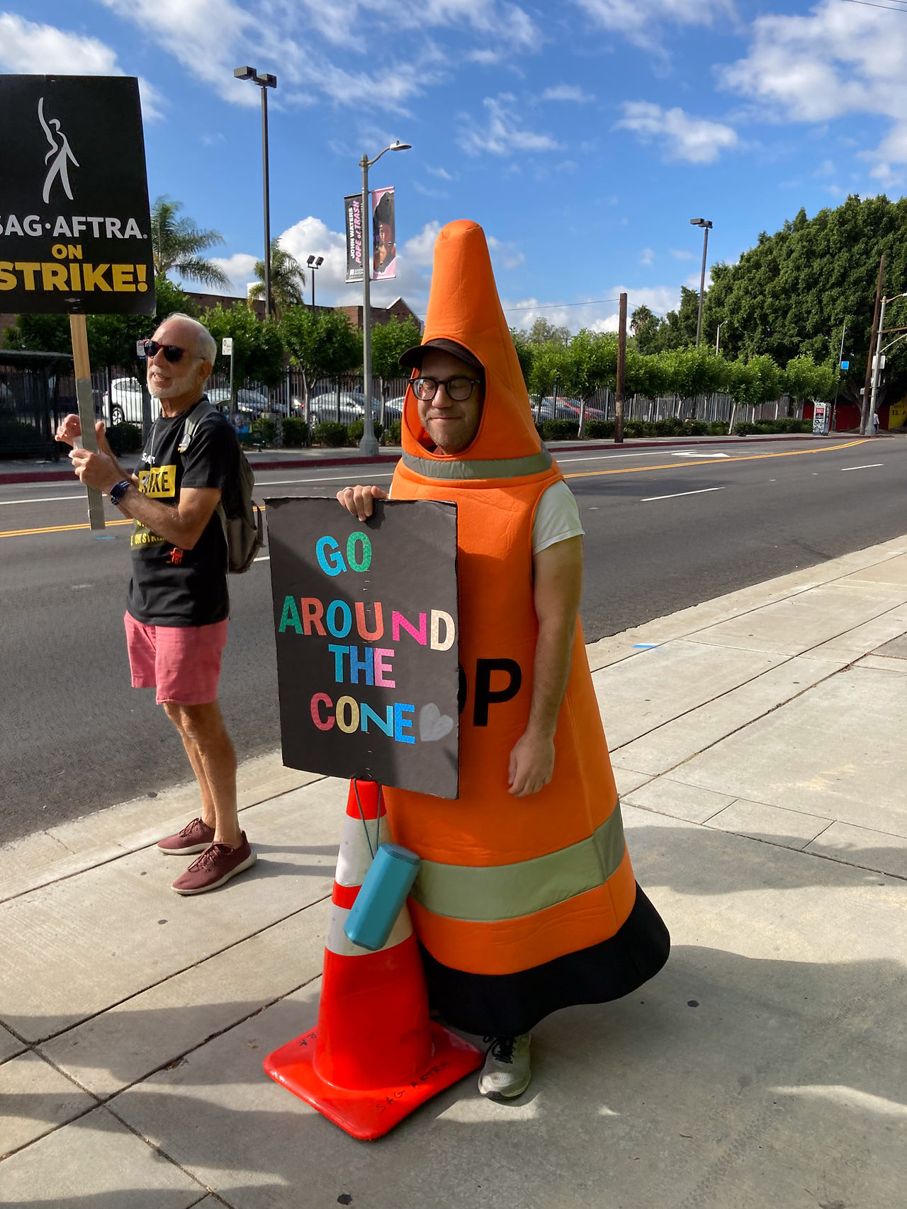 A strike captain, a man, dressed in an orange cone stands in line. The man dressed in a tall orange cone costume stands in line behind an orange traffic cone with a sign that reads "Go around the cone [heart]." The letters are colorful against a black backdrop. To his right, a man dressed in sunglasses, a black "SAG-AFTRA On Strike!" t-shirt and backpack, pink shorts and loafers holds a picket sign. Behind them is the street.