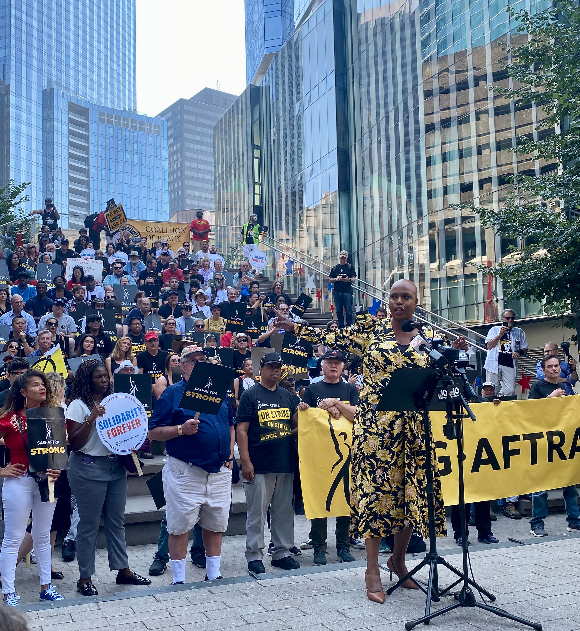 Representative Ayanna Pressley stands in front of a podium. Behind her on a set of bleachers or steps are SAG-AFTRA New England Local members. Protest signs can be seen throughout and further away, office buildings. 